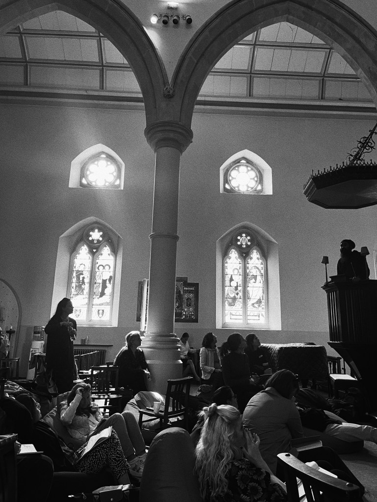 A monochromatic photograph featuring a group of people in a church, sitting on loungers and chairs and listening to speaker.